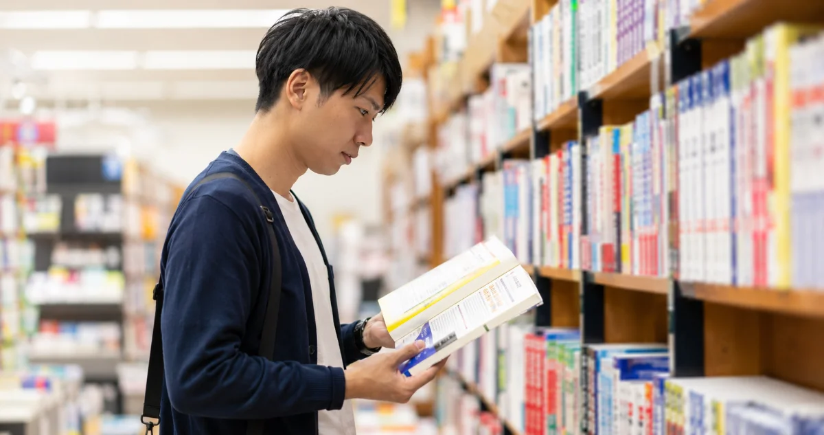 書店の資格書コーナーで試験対策の問題集を開いて見比べる男性。市販の模範解答は各社の解答例というイメージ