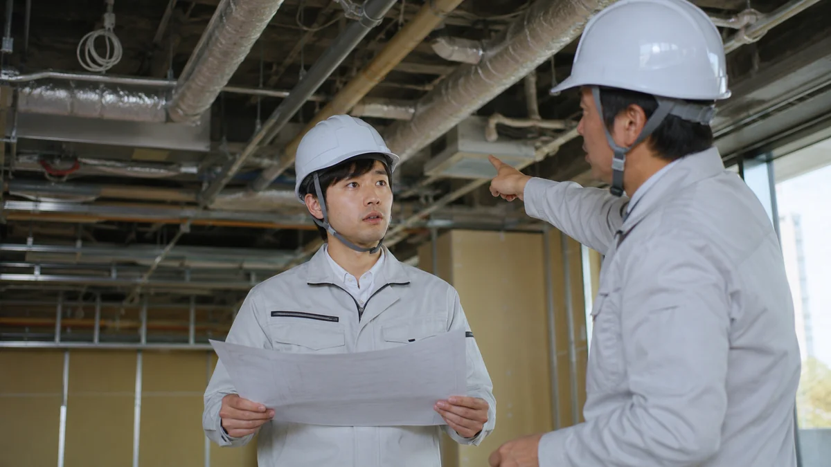 建設現場の天井裏で、図面を持つ若い設備施工管理者が元請けの担当者から指示を受けている様子