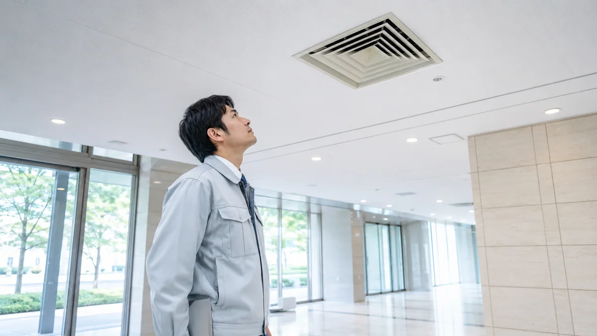オフィスビルの共用部で、天井の吹出口を見上げて点検している設備管理者